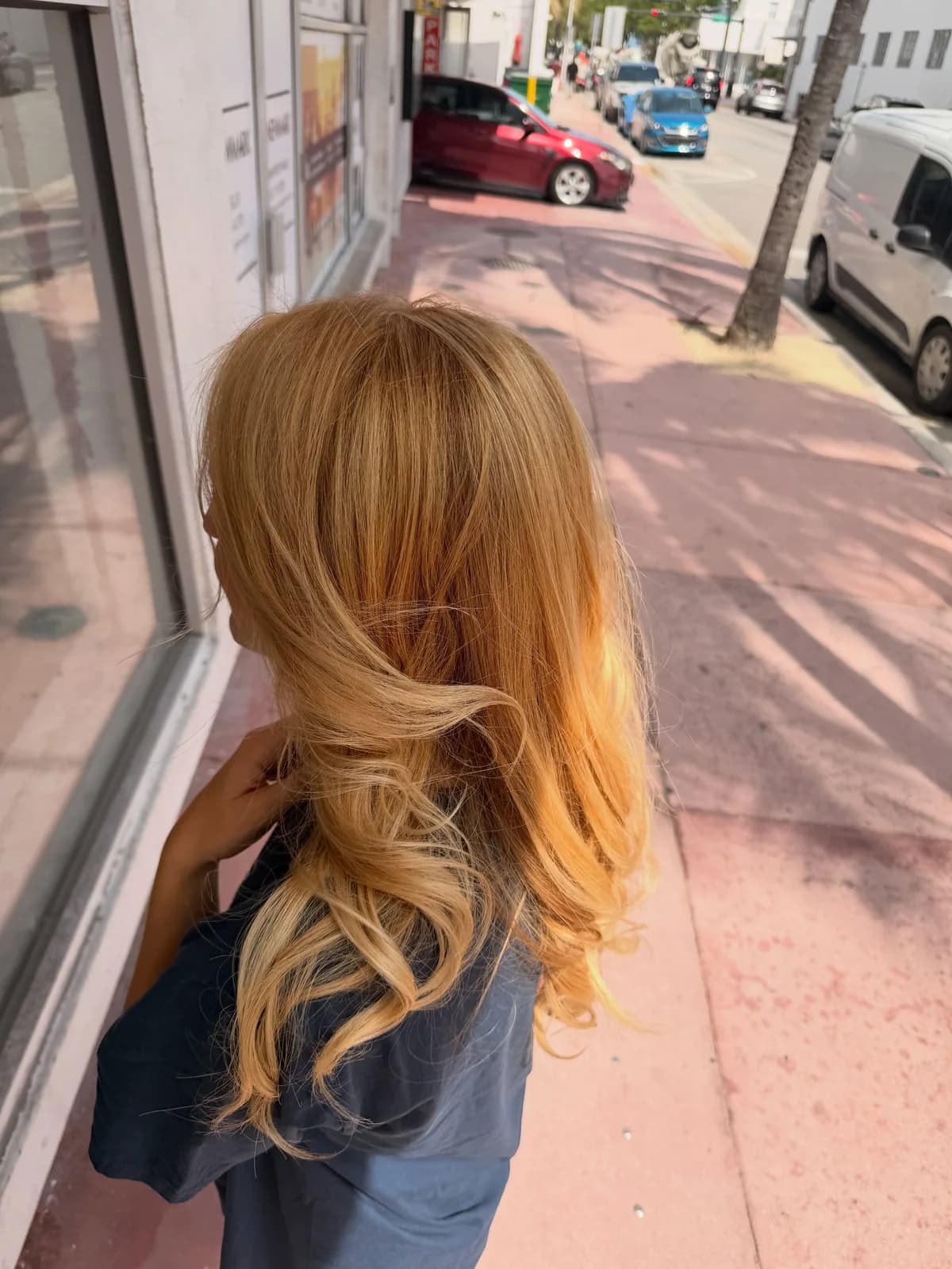 Golden blonde hair with blended roots and curled ends, photographed from behind on a sidewalk